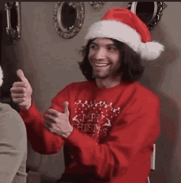 a man wearing a santa hat and a merry christmas sweater giving a thumbs up