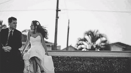 a black and white photo of a bride and groom holding hands while sitting on a wall .