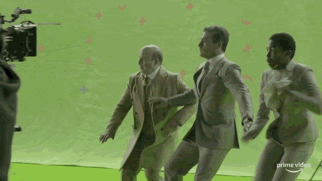 three men in suits are running on a green screen that says prime video on the bottom