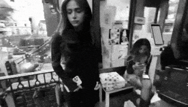 a black and white photo of a woman standing next to a woman sitting on a chair .