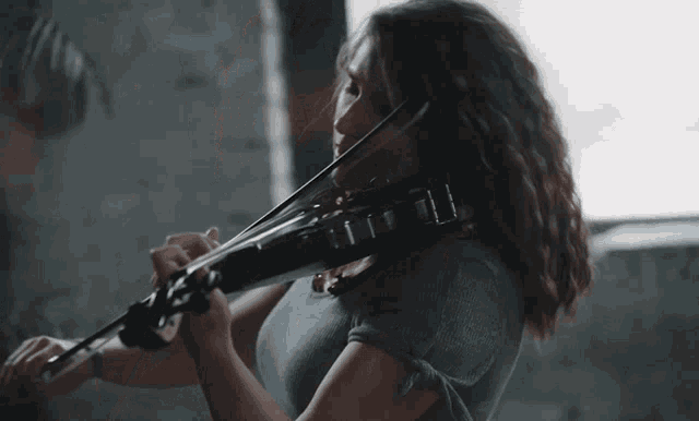 a woman in a blue shirt is playing a violin in front of a window
