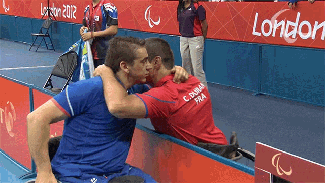 a man in a wheelchair is hugging another man in front of a london 2012 sign