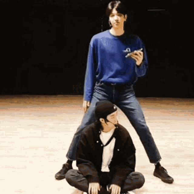 a man in a blue shirt is standing next to a man in a black hat sitting on the floor .