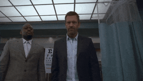 a man in a suit stands next to another man in front of an eye chart with the letter e on it