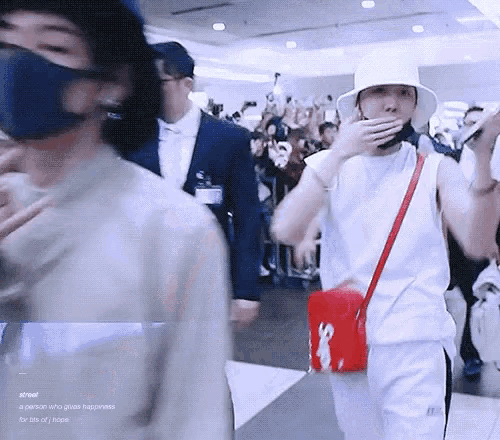 a man wearing a hat and a mask is covering his mouth with his hand while walking through an airport .