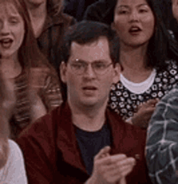 a man wearing glasses is sitting in a crowd of people clapping .