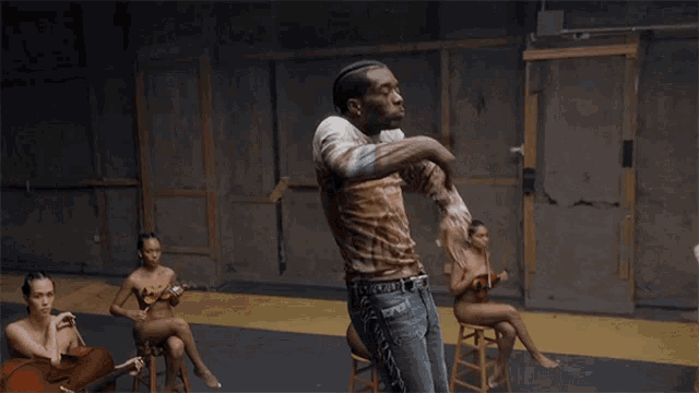a group of naked women sit on stools in a room with a man dancing
