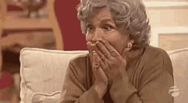 an elderly woman is sitting on a couch with her hands on her mouth .