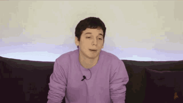 a young man in a purple shirt is sitting on a couch with a microphone in his mouth .