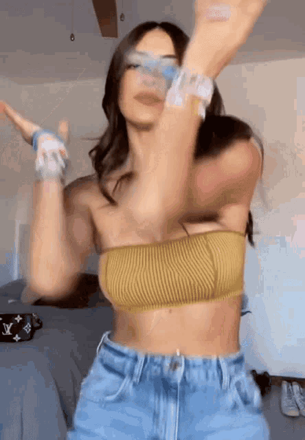 a woman in a yellow crop top and blue shorts is dancing in a bedroom .