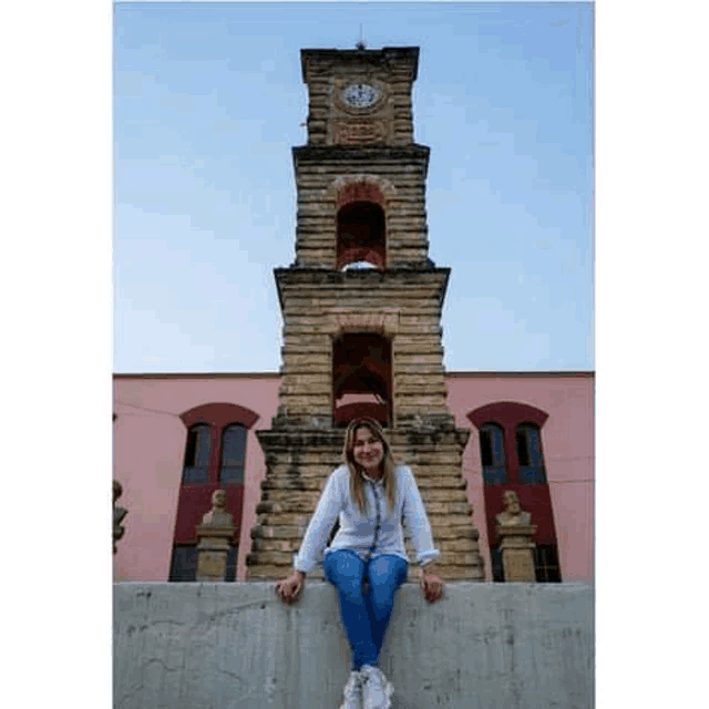 a woman is sitting on the edge of a wall in front of a clock tower .