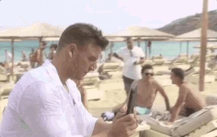 a man in a white shirt is standing on a beach holding a cell phone .