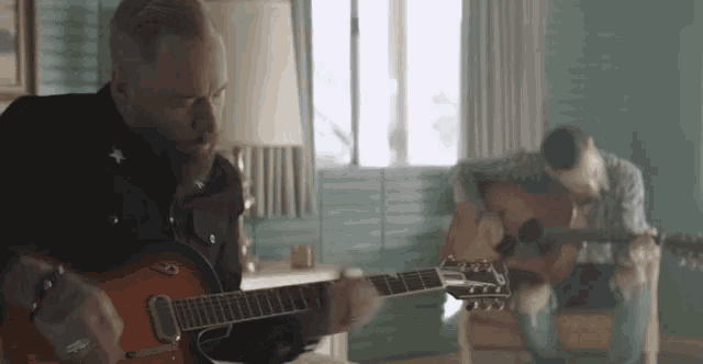 a man is playing a guitar while another man sits in a chair .