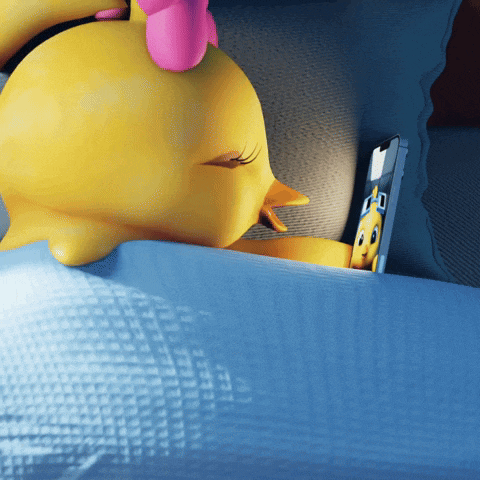 a yellow duck laying on a bed looking at a phone