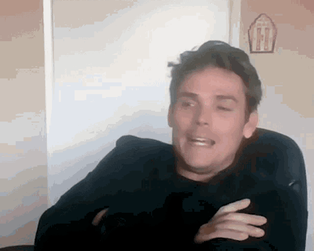 a man in a black sweater is sitting in a chair with his arms crossed and making a funny face .