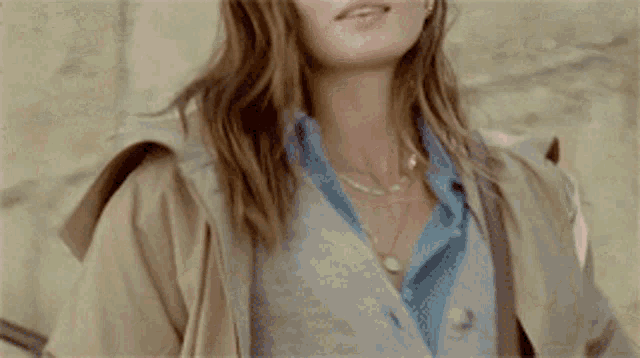 a woman in a tan coat and blue shirt is standing in front of a wall .