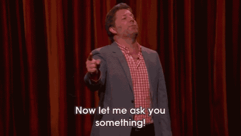 a man in a suit and red plaid shirt is standing on a stage and says `` now let me ask you something '' .