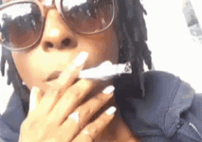 a woman with dreadlocks is smoking a cigarette while wearing sunglasses and a blue shirt .