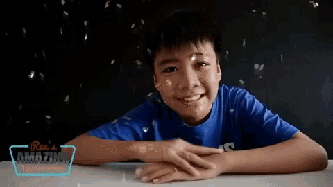a boy in a blue shirt is sitting at a table with confetti falling around him