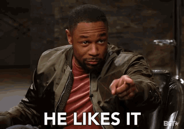 a man sitting in a chair pointing at the camera with the words he likes it below him
