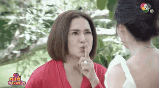 a woman in a red robe holds her finger to her mouth while talking to another woman in a white dress