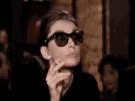 a woman wearing sunglasses is smoking a cigarette in a room .