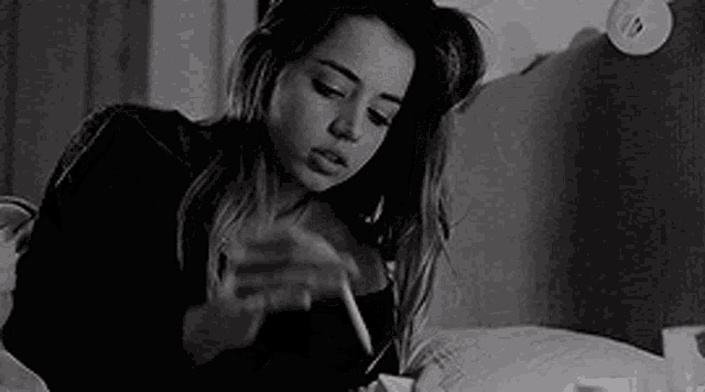 a black and white photo of a woman smoking a cigarette while sitting on a bed .