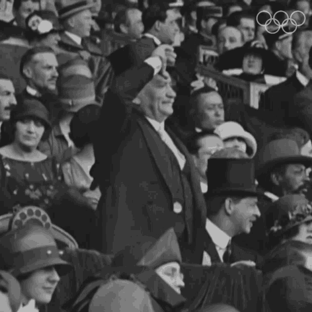 a man in a tuxedo stands in a crowd of people with the olympic rings behind him