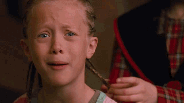 a little girl with braids is crying while a person braids her hair