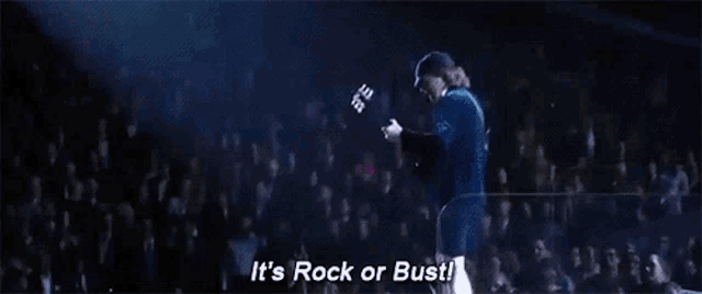 a man is standing on a stage in front of a crowd and says `` it 's rock or bust ! ''