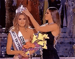 a woman is putting a crown on another woman who is holding a bouquet of flowers .