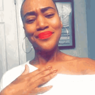 a woman with red lipstick and hoop earrings looks at the camera with her hand on her chest