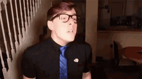a man wearing glasses , a black shirt and a blue tie is standing in front of a staircase .