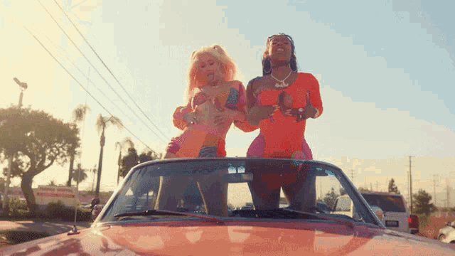 two women standing on top of a red car with one wearing a necklace that says ' barbie ' on it
