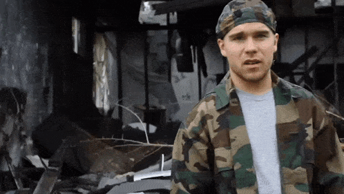 a man wearing a camouflage jacket and a headband stands in front of a destroyed building