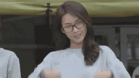 a woman wearing glasses and a light blue sweater is standing in front of a tent .