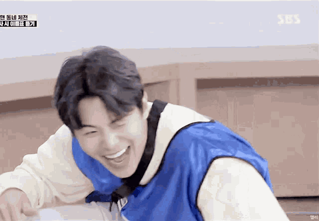 a young man wearing a blue vest is laughing in front of a sbs logo