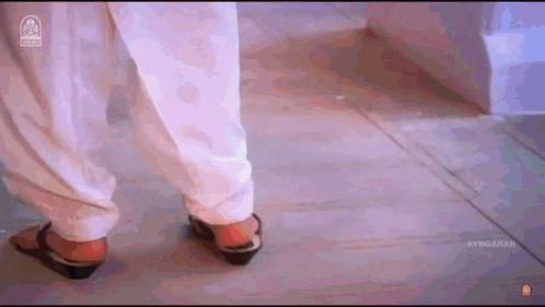a woman wearing white pants and black shoes is walking on a wooden floor .