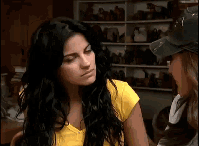 a woman with long black hair is wearing a yellow shirt and a baseball cap and looking at the camera .