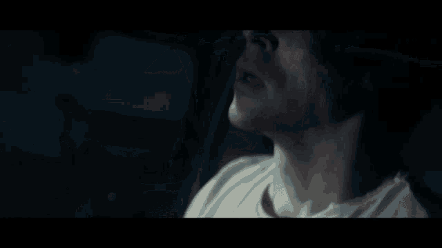 a man in a white shirt is sitting in a dark room with his hand on his ear .