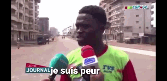 a man wearing a tvs shirt is talking into a microphone on a street