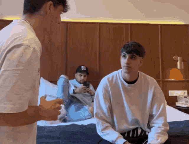 a man sitting on a bed talking to another man while another man sits on the floor