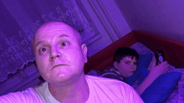 a man in a white shirt looks at the camera while a boy plays a video game in the background