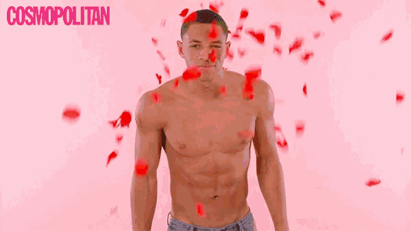a shirtless man is standing in front of a pink background with roses falling on him .