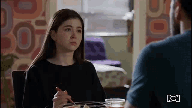 a girl is sitting at a table with a fork in her hand while talking to a man .