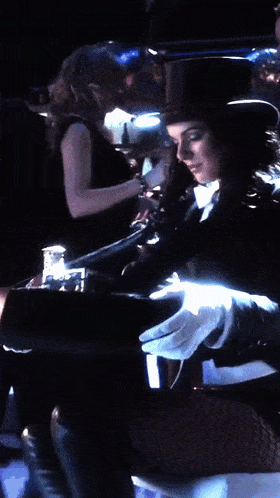 a woman wearing a top hat and gloves is sitting in a dark room