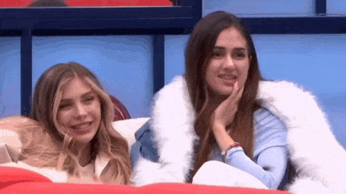 two women are sitting next to each other on a couch and one is wearing a white fur coat .