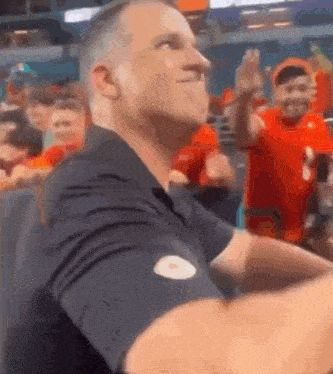 a man in a black shirt is sitting in a stadium with other people .