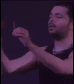 a man wearing sunglasses and a black tank top is singing into a microphone in a dark room .
