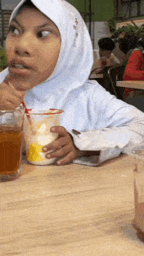 a girl in a hijab is sitting at a table holding a drink